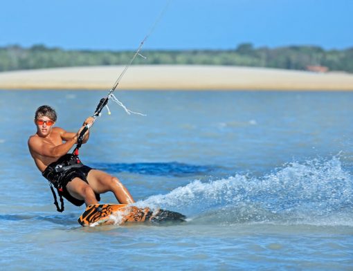Kitesurfista de pé na prancha cortando a água e levantando espuma, preso à vela pelo trapézio