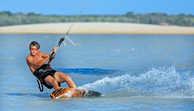 Kitesurfista de pé na prancha cortando a água e levantando espuma, preso à vela pelo trapézio
