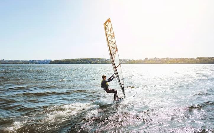 Homem se deslocando sobre as águas de uma praia com um windsurf.