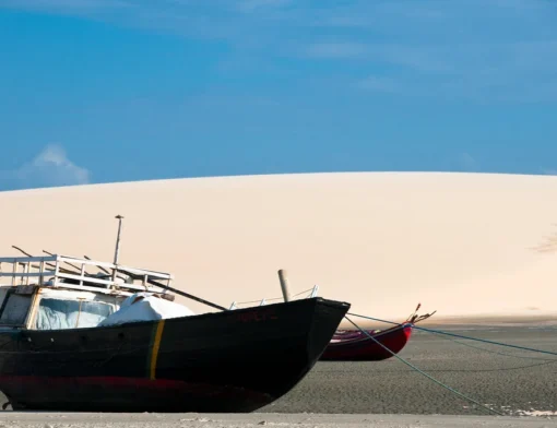 Barco de pesca de casco escuro encalhado na areia úmida, com uma grande duna branca ao fundo e céu azul