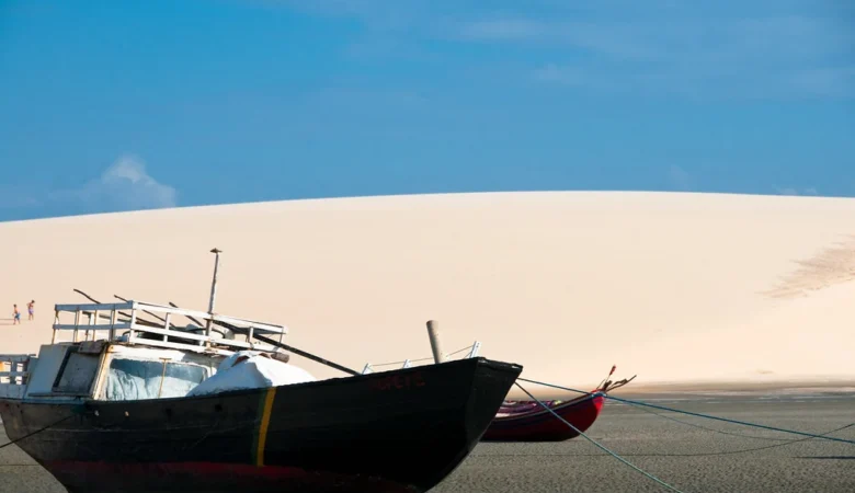 Barco de pesca de casco escuro encalhado na areia úmida, com uma grande duna branca ao fundo e céu azul