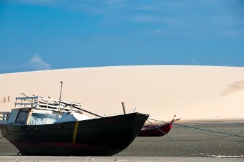 Barco de madeira encalhado na areia com grande duna branca ao fundo, nos Lençóis Maranhenses