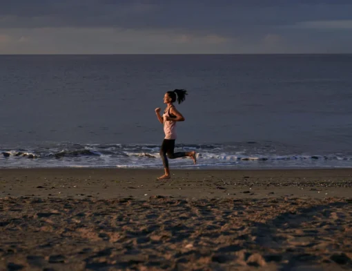 Mulher correndo descalça na areia da praia ao entardecer, com o mar ao fundo