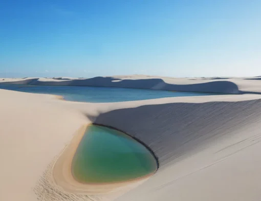 Lagoa de água verde no pé de uma duna branca, com outra lagoa azul e mais dunas ao fundo, sob céu azul