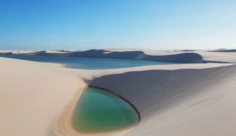 Lagoa de água verde no pé de uma duna branca, com outra lagoa azul e mais dunas ao fundo, sob céu azul