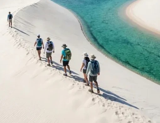 Grupo de caminhantes atravessando o cume de uma duna de areia branca, com lagoa de água cristalina em tons de verde e azul à direita