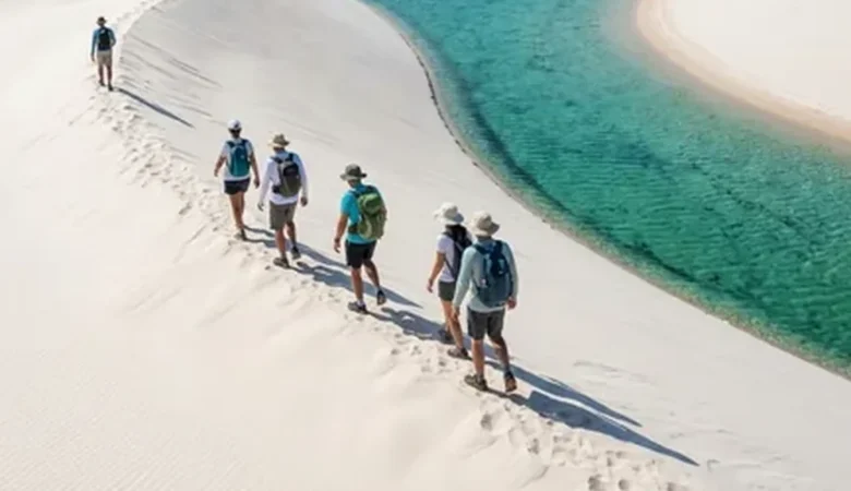 Grupo de caminhantes atravessando o cume de uma duna de areia branca, com lagoa de água cristalina em tons de verde e azul à direita