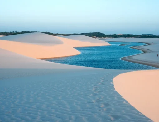 Lagoa de água azul serpenteando entre dunas brancas iluminadas pelo sol baixo, com pessoas caminhando na margem