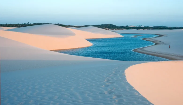 Lagoa de água azul serpenteando entre dunas brancas iluminadas pelo sol baixo, com pessoas caminhando na margem