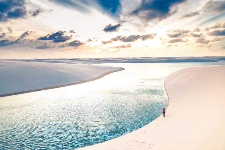 Lagoa de águas azuis entre dunas brancas dos Lençóis Maranhenses ao entardecer, com uma pessoa caminhando na areia