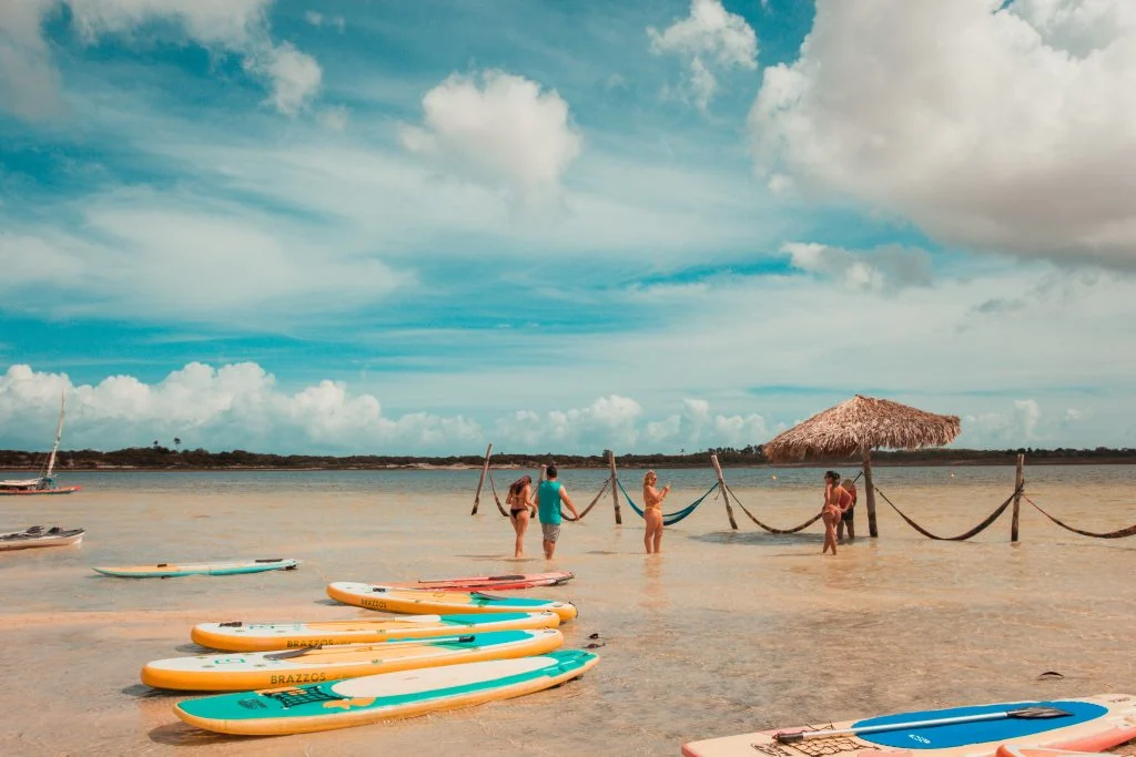 Pranchas de stand up paddle na margem de uma lagoa rasa, com redes e guarda-sol de palha dentro da água
