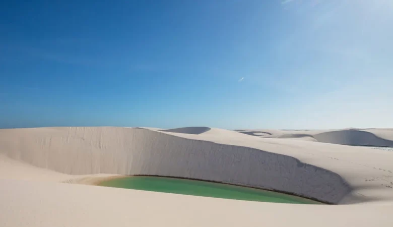 Lagoa de água verde encaixada entre dunas de areia branca, sob céu azul limpo