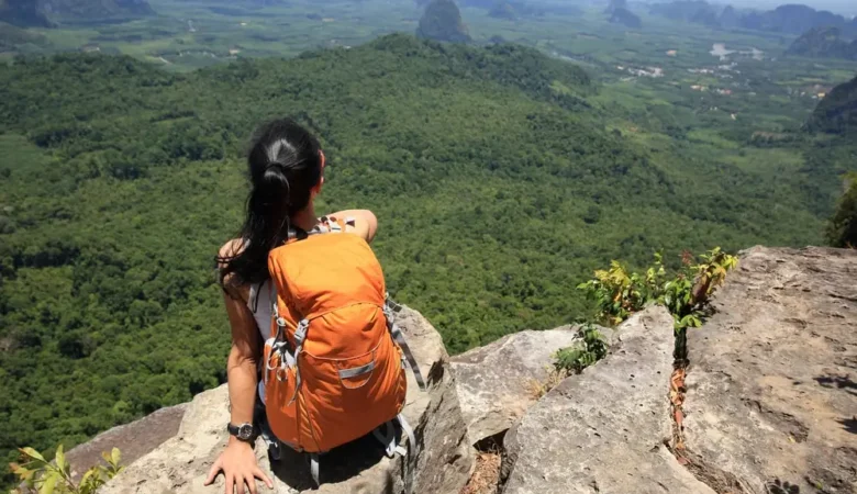 Mochileira sentada em um rochedo observando um vale coberto de floresta, com morros ao fundo