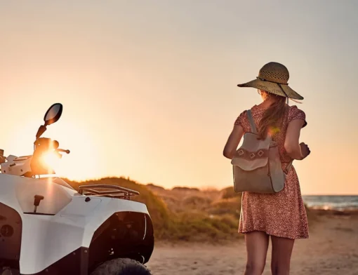 Mulher de chapéu de palha e mochila ao lado de um quadriciclo branco, ao pôr do sol perto do mar