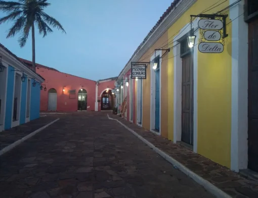 Rua de pedra do Porto das Barcas, em Parnaíba, entre casarões coloridos com lampiões acesos e placas de lojas, ao entardecer