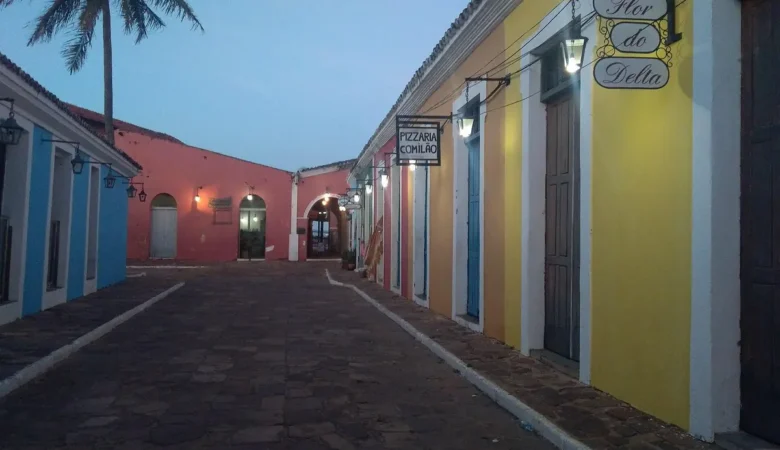 Rua de pedra do Porto das Barcas, em Parnaíba, entre casarões coloridos com lampiões acesos e placas de lojas, ao entardecer