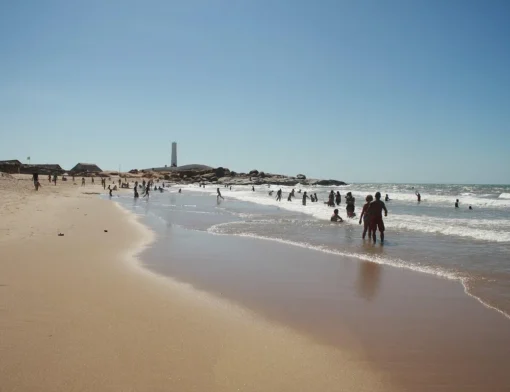 Banhistas no mar da Praia da Pedra do Sal, em Parnaíba, com rochedos de granito e o farol branco ao fundo