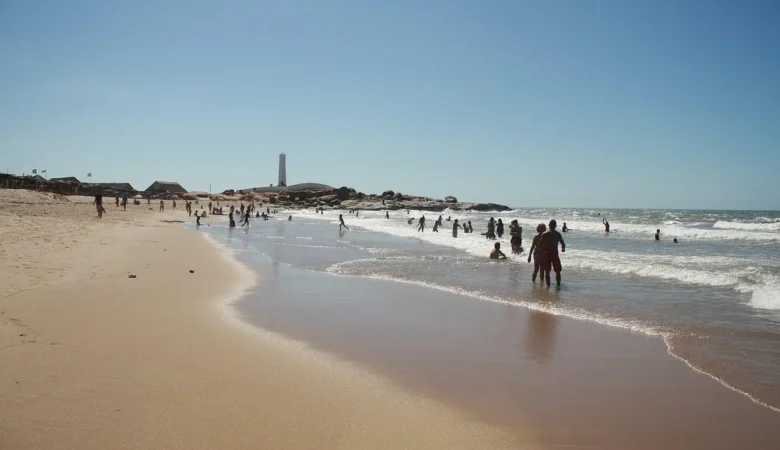 Banhistas no mar da Praia da Pedra do Sal, em Parnaíba, com rochedos de granito e o farol branco ao fundo