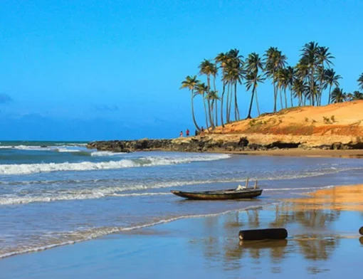 Praia com mar batendo na areia, canoa encalhada, falésia alaranjada e fileira de coqueiros ao fundo
