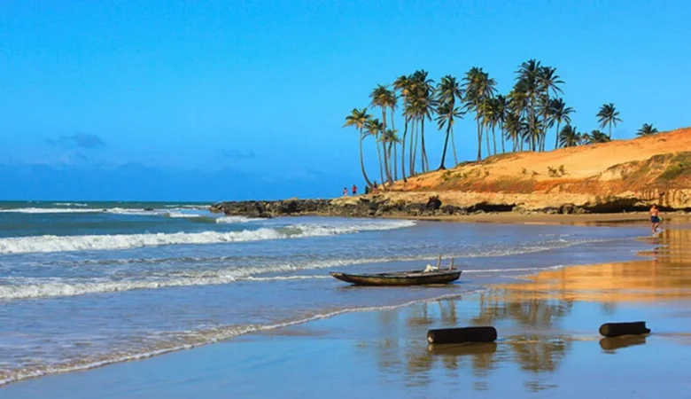 Praia com mar batendo na areia, canoa encalhada, falésia alaranjada e fileira de coqueiros ao fundo