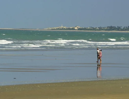 Casal caminhando na areia molhada da Praia Peito de Moça, com ondas, dunas, coqueiros e o farol da Ponta do Itaqui ao fundo