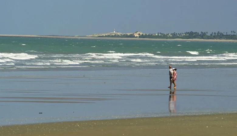 Casal caminhando na areia molhada da Praia Peito de Moça, com ondas, dunas, coqueiros e o farol da Ponta do Itaqui ao fundo