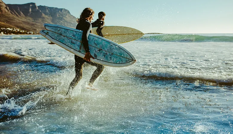 Dois surfistas de roupa de neoprene correndo para o mar com pranchas, com montanhas ao fundo