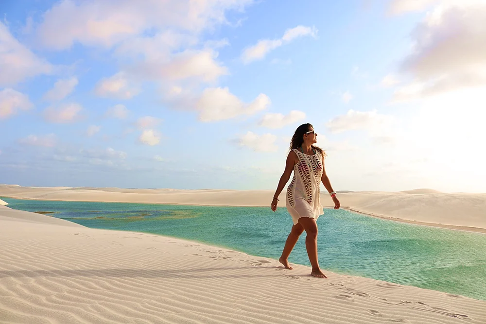 Visitante caminhando descalça no alto de uma duna branca diante de lagoa de água esverdeada nos Lençóis Maranhenses